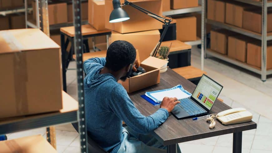 homem, sentado em uma mesa, na frente de um notebook, dentro de um estoque, analisando um produto na caix