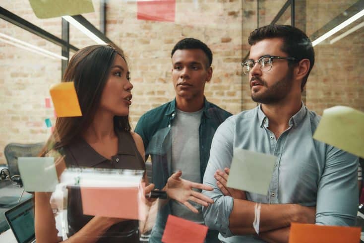 Dois homens e uma mulher conversando um frente a um espelho com post-its.